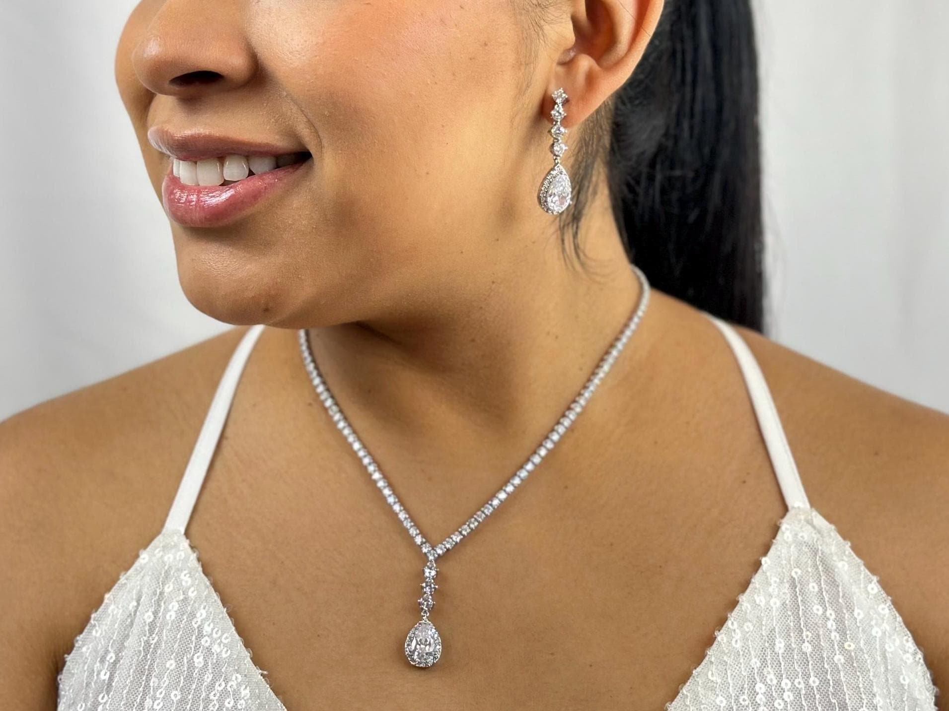 Close-up of a model’s face and neck showcasing elegant dainty diamond sparkly teardrop earrings and a matching necklace with a pendant. The model is smiling, with her face turned to the left, highlighting the beauty of the sparkly diamond jewelry set