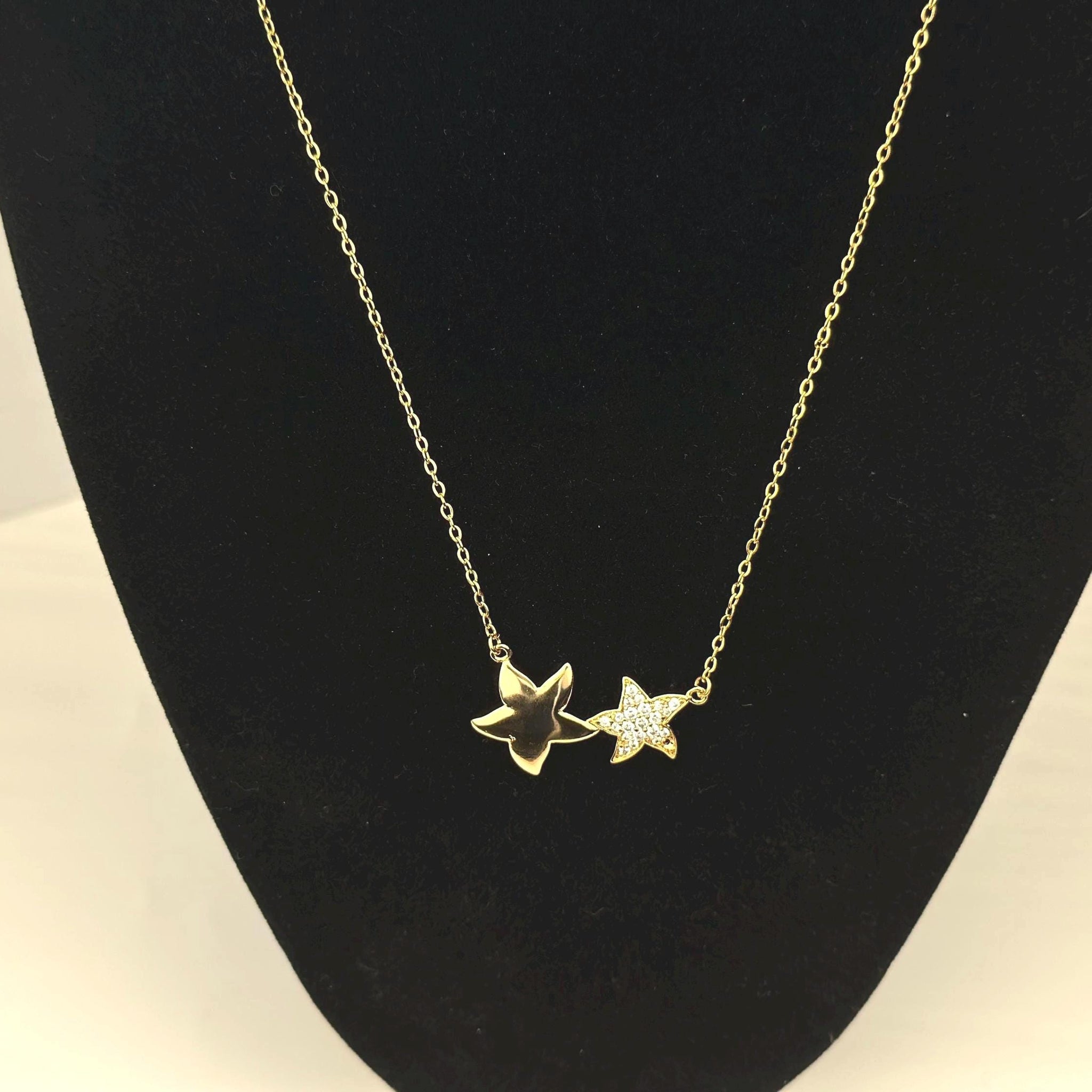 18K gold filled necklace with double star pendant. The stars are of different sizes sitting side by side. The larger star is a smooth gold surface, the second smaller star sparkling gemstones.The necklace hung on a black prop to emphasize the beauty of the necklace.