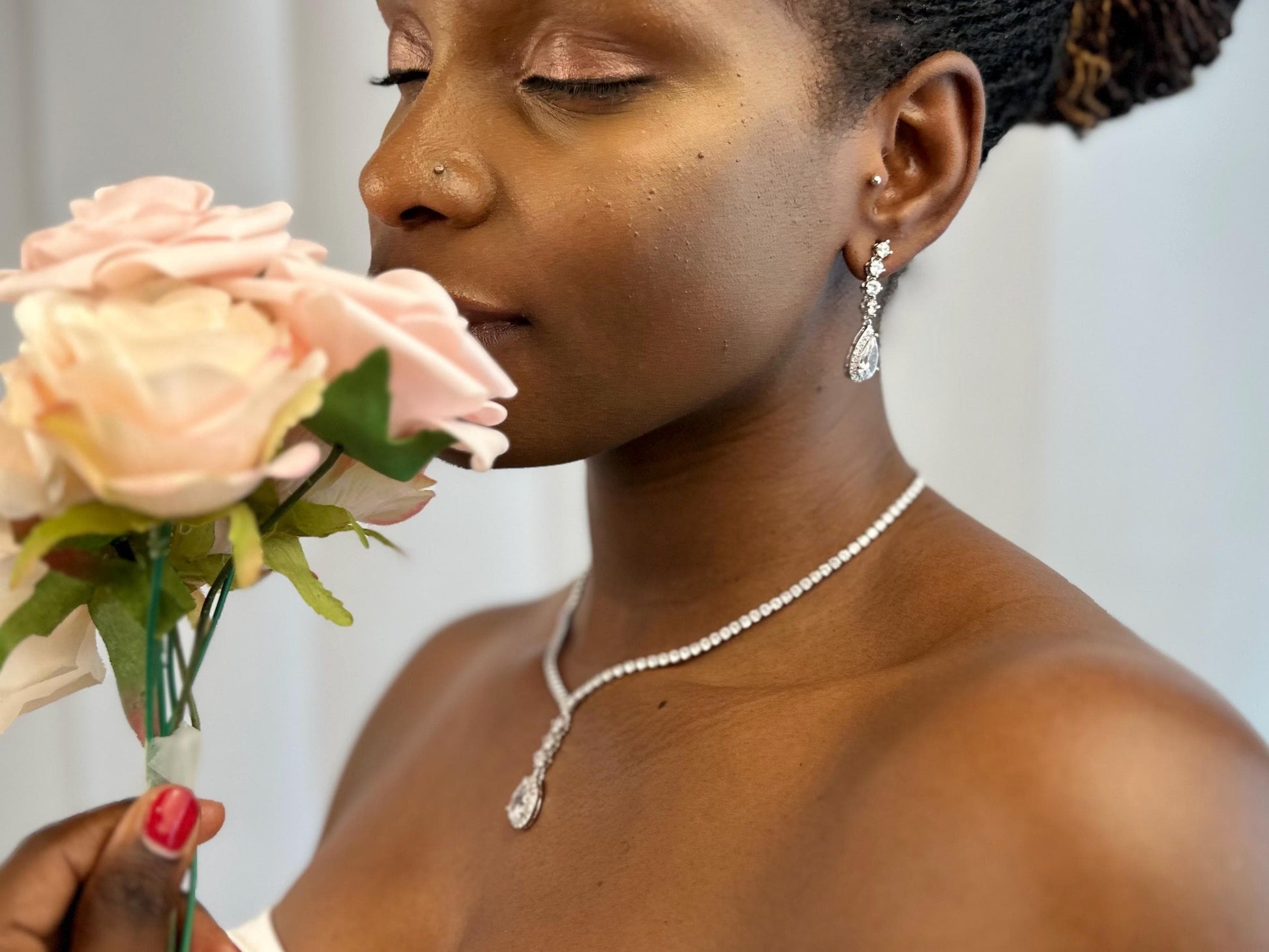 Close-up of a bride’s face and neck showcasing elegant dainty diamond sparkly teardrop earrings and a matching necklace with a pendant. The bride is enjoying the aroma of a bouquet, highlighting the magical sparkle and beauty of the jewelry set