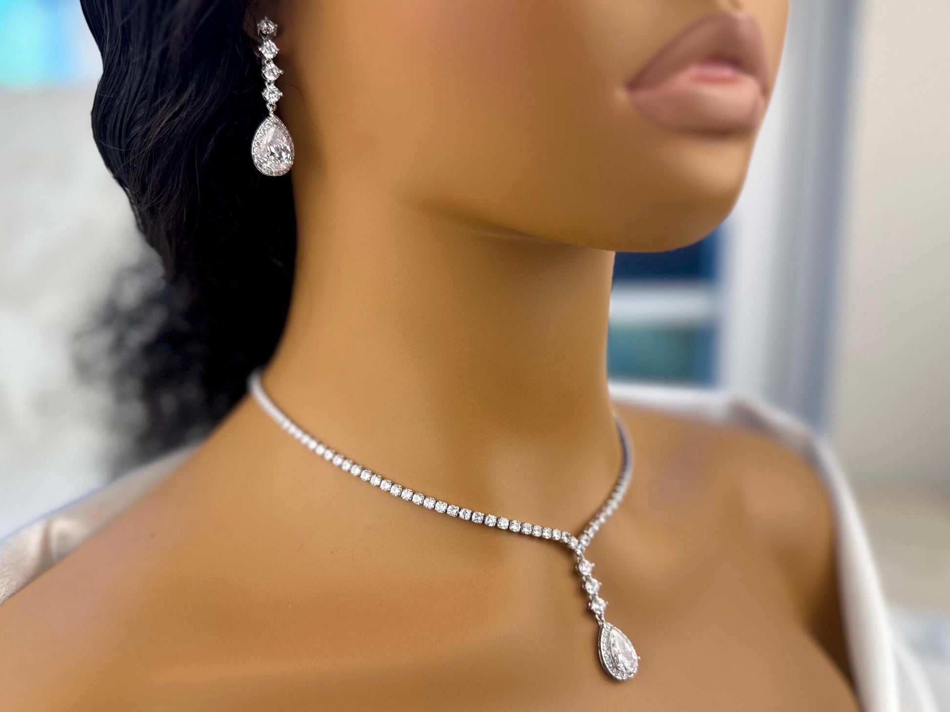 Close-up of a mannequin’s lower face and neck showcasing elegant dainty diamond sparkly silver bridal earrings and a matching necklace with a pendant. The jewelry is set against a blurred blue background, highlighting the sparkle of the diamonds