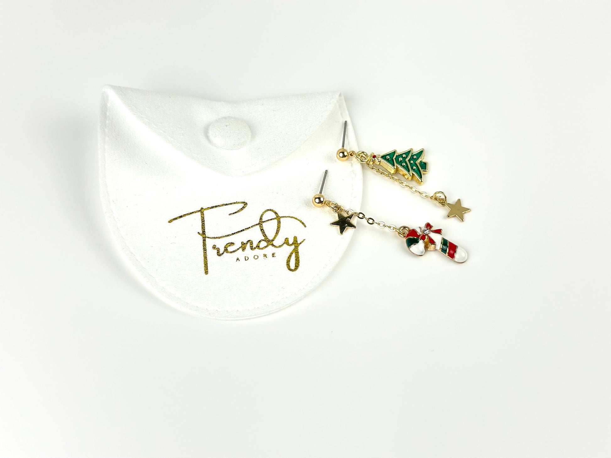 Next to a white jewelry Pouch with TrendyAdore branded in Gold, are a pair of mismatched earring: one with green christmas tree and gold star charm hanging down in a small chain, and the other is a red, green and white candy cane and gold star charm.