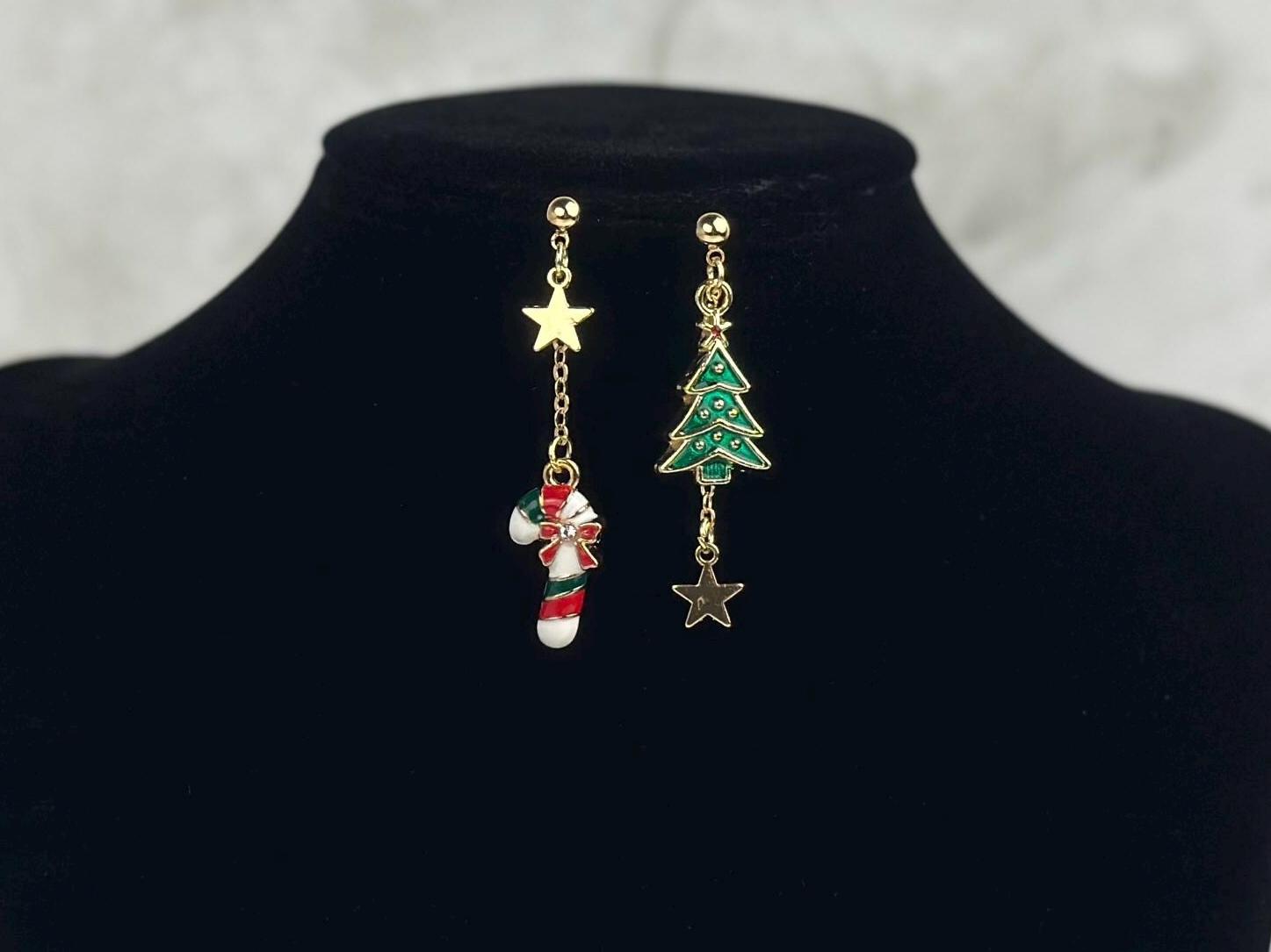 Attached to a black jewelry prop are a pair of mismatched earrings: one with green christmas tree and gold star charm hanging down in a small chain, and the other is a red, green and white candy cane and gold star charm.