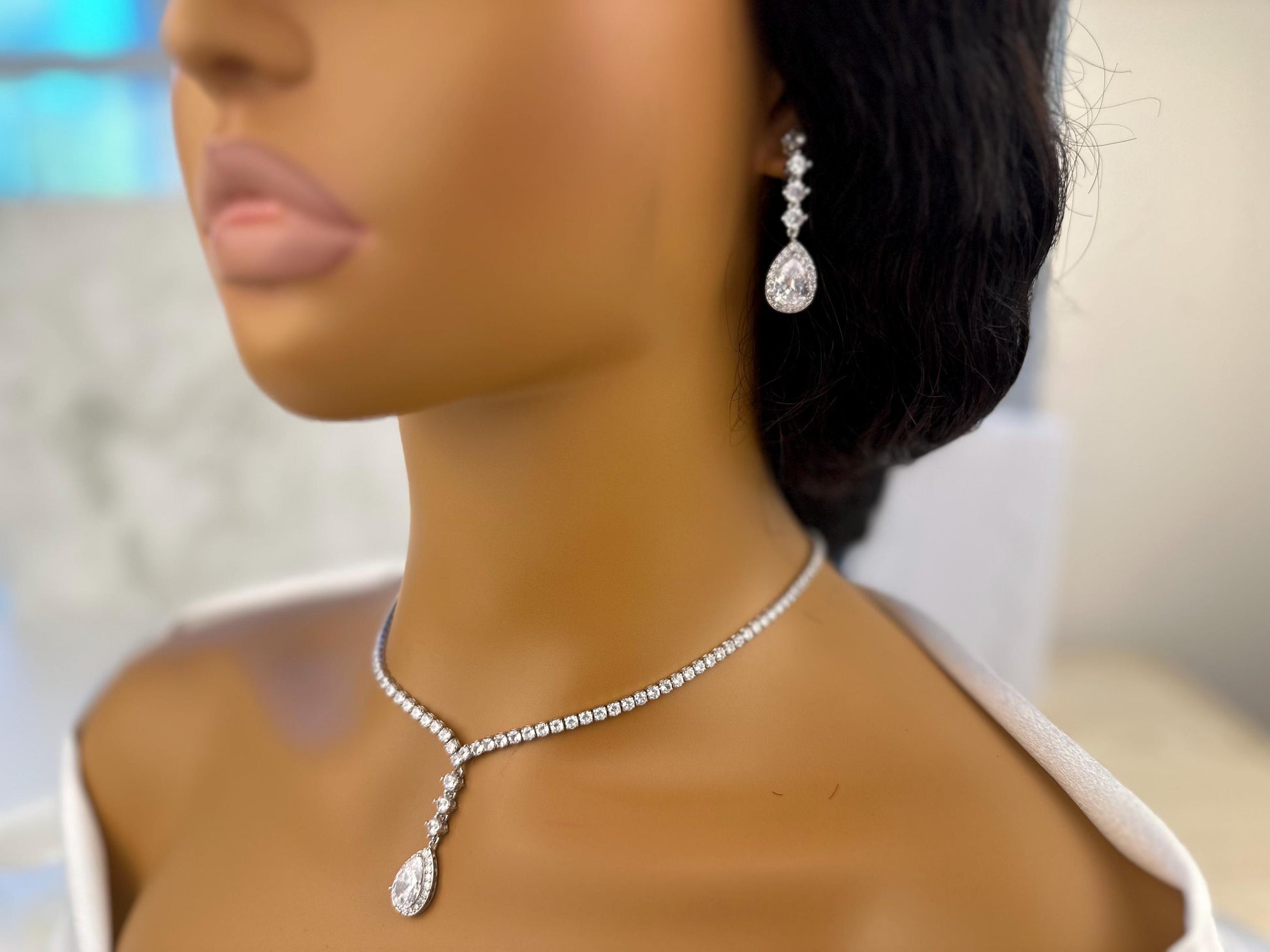 Close-up of a mannequin’s lower face and neck showcasing elegant dainty diamond sparkly teardrop earrings and a matching necklace with a pendant. The jewelry is set against a blurred blue background, highlighting the sparkle of the diamonds